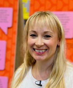 Victoria Newton, with long, blonde hair, wearing a white and multi-coloured t-shirt, standing in front of bright orange, pink and green notice boards