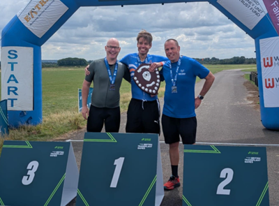 Three men in sportswear, one holding a shield, standing behind first, second and third place podiums in a field
