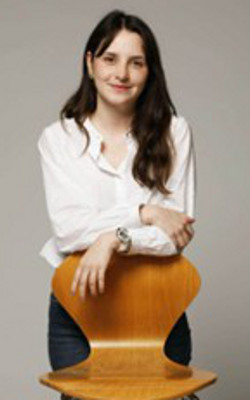 Betriz Ceni Lopes, with long dark hair, wearing a white blouse and blue jeans, standing behind a wooden chair with her hands draped over the back of the chair