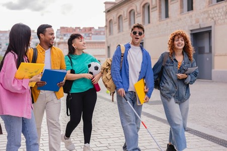 A group of young men and women, wearing brightly coloured clothes and holding folders, walking along a path. One young man is holding a white cane