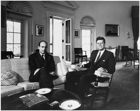 A black and white photo of Valery Giscard d'Estaing sitting on a sofa next to President John F Kennedy, who is sitting on a chair