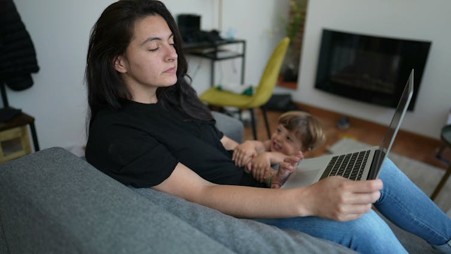 A lady wearing a black t-shirt and jeans, sitting on a sofa holding a laptop, with a young child on the floor next to her, holding on to her arm