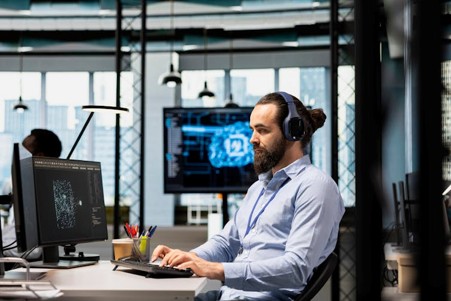 A man wearing a blue shirt and headphones, sitting at a desk with a monitor on it, typing on a keyboard