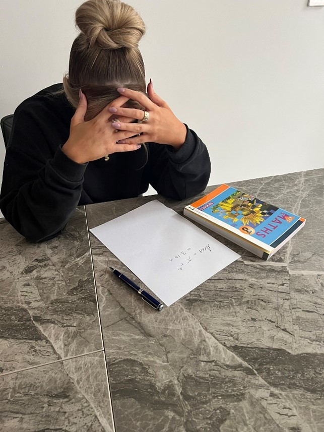 A lady sitting at a table with her head in her hands, and a math book and a paper with a maths calculation on it on the table
