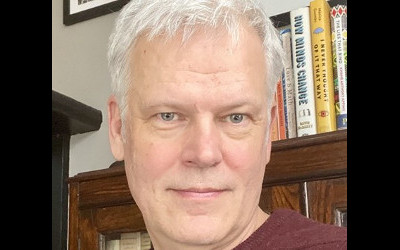 Paul Piwek, with short, grey hair, wearing a maroon-coloured top, in front of a bookshelf