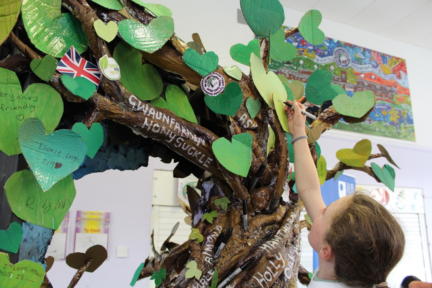 A sculpture of a tree with green paper leaves on it, with somebody reaching up to write on one of the leaves