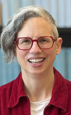 Rebecca L Jones, smiling at the camera and wearing red glasses, a white t-shirt and a red, collared shirt