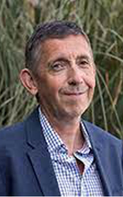 Robin Mellors-Bourne, wearing a blue striped shirt and blue jacket, standing in front of greenery