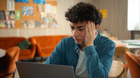 A teenage boy, with dark, curly hair, wearing a white t-shit and a blue denim shirt, sitting with his face resting on his hand and looking a a laptop. There are orange sofas and chairs in the background.