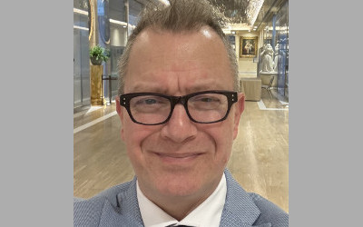 Theo Papaioannou, wearing a pair of glasses, a white, collared shirt and a grey jacket