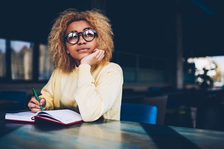 Woman wearing glasses, sitting at a desk with a notebook and pen, looking into the distance