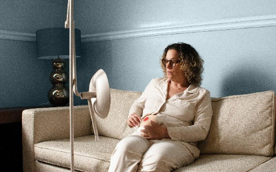 A woman wearing cream pyjamas, sitting on a cream sofa, wearing a stoma with a stoma product on a stand in front of her