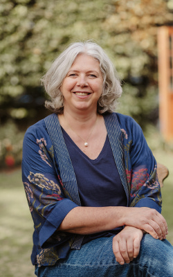 Victoria Nicholas, wearing a blue top and jeans, sitting with her hands crossed in her lap, with a background of trees and grass behind her