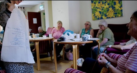 A group of women sitting around tables looking at a lady holding a white piece of fabric with wording on it