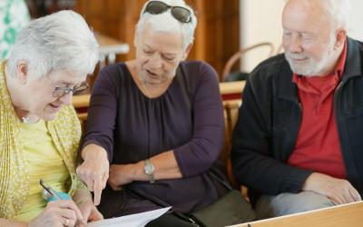 Three elderly people, two woman and a man, sitting together and looking at one of the women who is writing on a piece of paper
