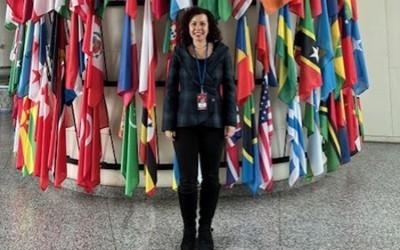 Dr Alessandra Marino, dressed in black top and trousers, standing in front of lots of multi-coloured flags
