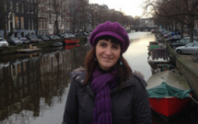 Angeliki Lymberopoulou, wearing a purple hat and scarf and a black coat, standing next to a river
