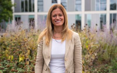 Anna De Liddo, with long blonde hair, wearing a white top and a beige jacket, standing in front of glass windows of a building and bushes