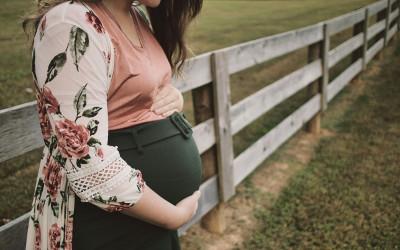 A pregnant woman, standing in front of a wooden fence and wearing a pink top, a floral jacket and green trousers, cradling her baby bump