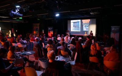 A group of people sitting round tables watching a lady standing behind a lecturn with presentation slides on a screen behind her