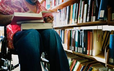 A disabled person, sitting in a wheelchair in a library, with several books in their lap