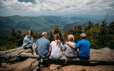 A group of five men and women sitting on a log overlooking a view of trees and mountains