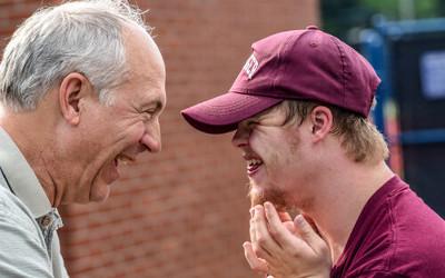 A man with grey hair and a grey shirt talking to a Down syndrome man wearing a maroon baseball cap and a maroon t-shirt