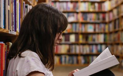 A young girl, with long, dark hair and wearing a white top and glasses, sitting in a library reading a book