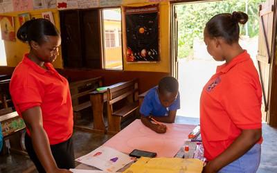 Two African teachers wearing red tops in a classroom with a young school boy with  pencil in his hand drawing on a piece of paper