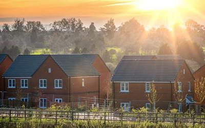 Several red-brick houses, with grey roofs and white window frames, with green grass fields, trees and a sunset in the background