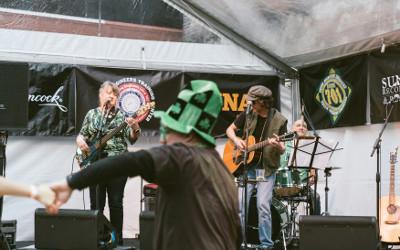 A band with two people on guitars and one on drums playing on a stage, with a person wearing a green hat with shamrocks on it dancing in front of them
