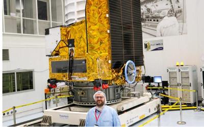 Jesper Skottfelt, wearing blue overalls and a plastic hat, standing in front of space equipment in a lab