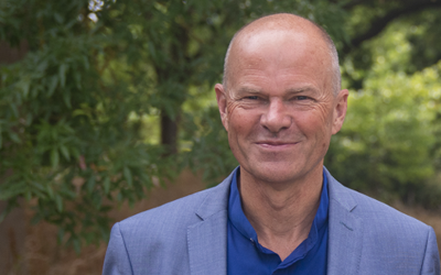 Head shot of a man wearing a blue shirt and jacket, with trees in the background