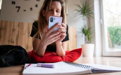 A lady holding a mobile phone with her arms resting on a red cushion, with an open notebook and pen on a table on front of her. Gajus/Shutterstock