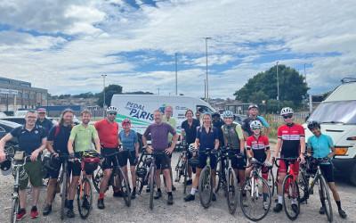 A group of cyclists posing for the camera, with white vans and vehicles on the background