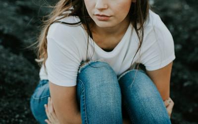 A girl with long dark hair, wearing a white t-shirt and jeans, sitting and hugging her knees