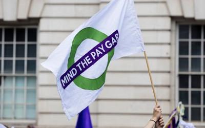 Somebody bearing a white flag, with a green circle on it and the words 'Mind the pay gap' on it