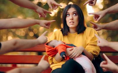 A mother sitting on an orange bench, with a baby in her arms, with lots people pointing fingers at her