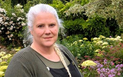 Naomi Miller, with blonde hair in a plait, wearing a green top, standing in front of bushes and yellow and pink flowers