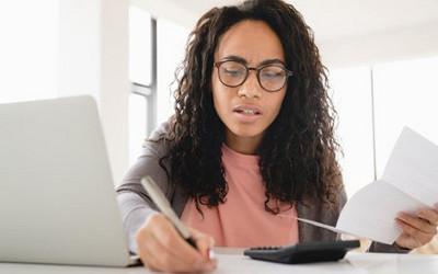 Woman writing with a pen and holding a piece of paper, with a laptop and calculator on the table