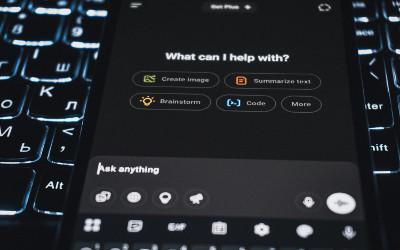 A smartphone with the words "What ca I help with?" on it, sitting on top of a keyboard
