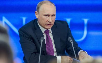 Vladimir Putin, wearing a suit and a purple tie, sitting in front of microphones
