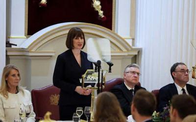 Rachel Reeves, standing in front of a microphone, surrounded by a group of men and women sitting around her 