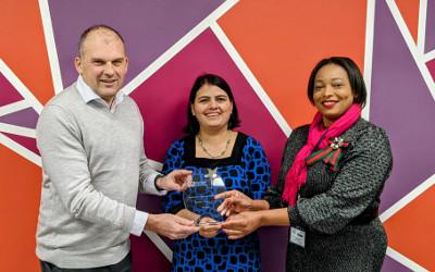 A man in a grey jumper standing on the left of two women, one with dark hair and wearing a blue top and the other with dark hair, wearing a black and white dress and a pink scarf, all holding a glass trophy
