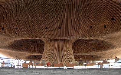 A tall, mushroom-shaped wooden building structure, with tables, seats and people underneath it