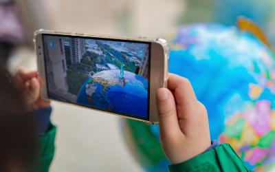 Child's hands holding a mobile phone with an image of a globe, a statue and buildings on it, with a globe in the background