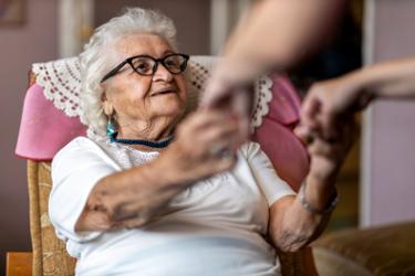 Elderly woman, sitting in a chair, reaching her hands out to a carer