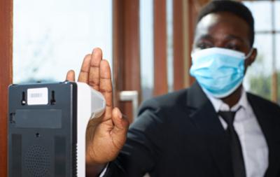 A black man wearing a suit and an anti-virus mask is using his hand to scan his body temperature