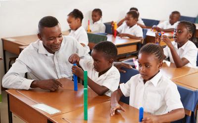 School children sitting at desks playing with coloured bricks, with a teacher helping them