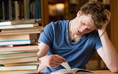 Man wearing a blue t-shirt, with his head in his hand, reading a book, with a pile of books on the table next to  him